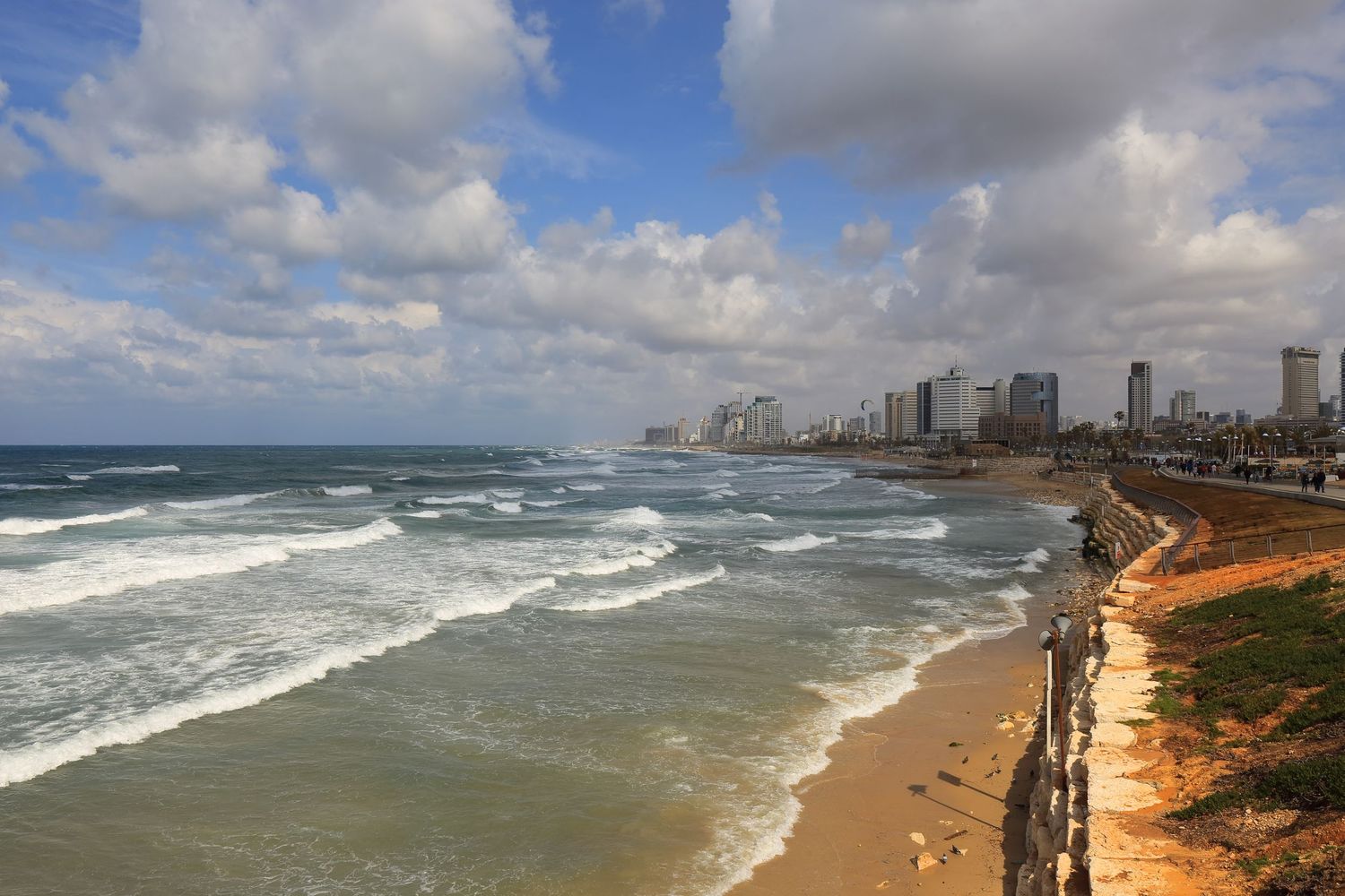 Scenic coastal view of Tel Aviv's beachfront with crashing waves and high-rise buildings under cloudy skies.