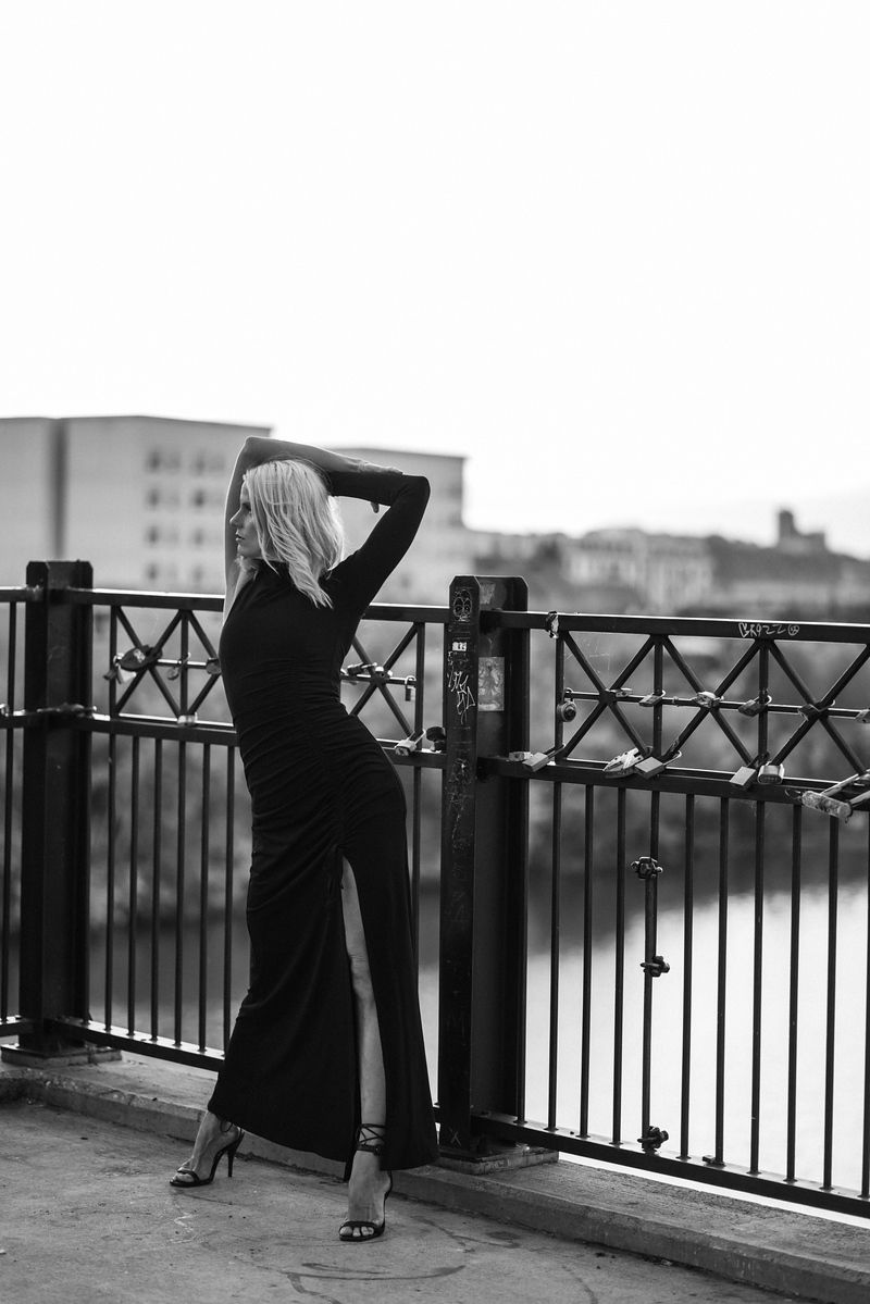 Black and white portrait of figure in long dress posing against riverside railing.