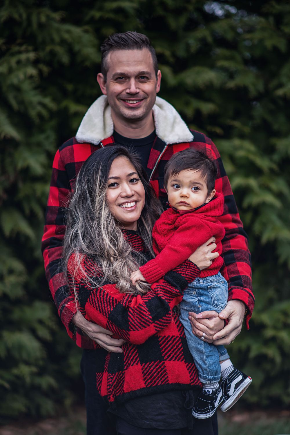 A family dressed in red and black buffalo plaid poses together outdoors against a natural green background.