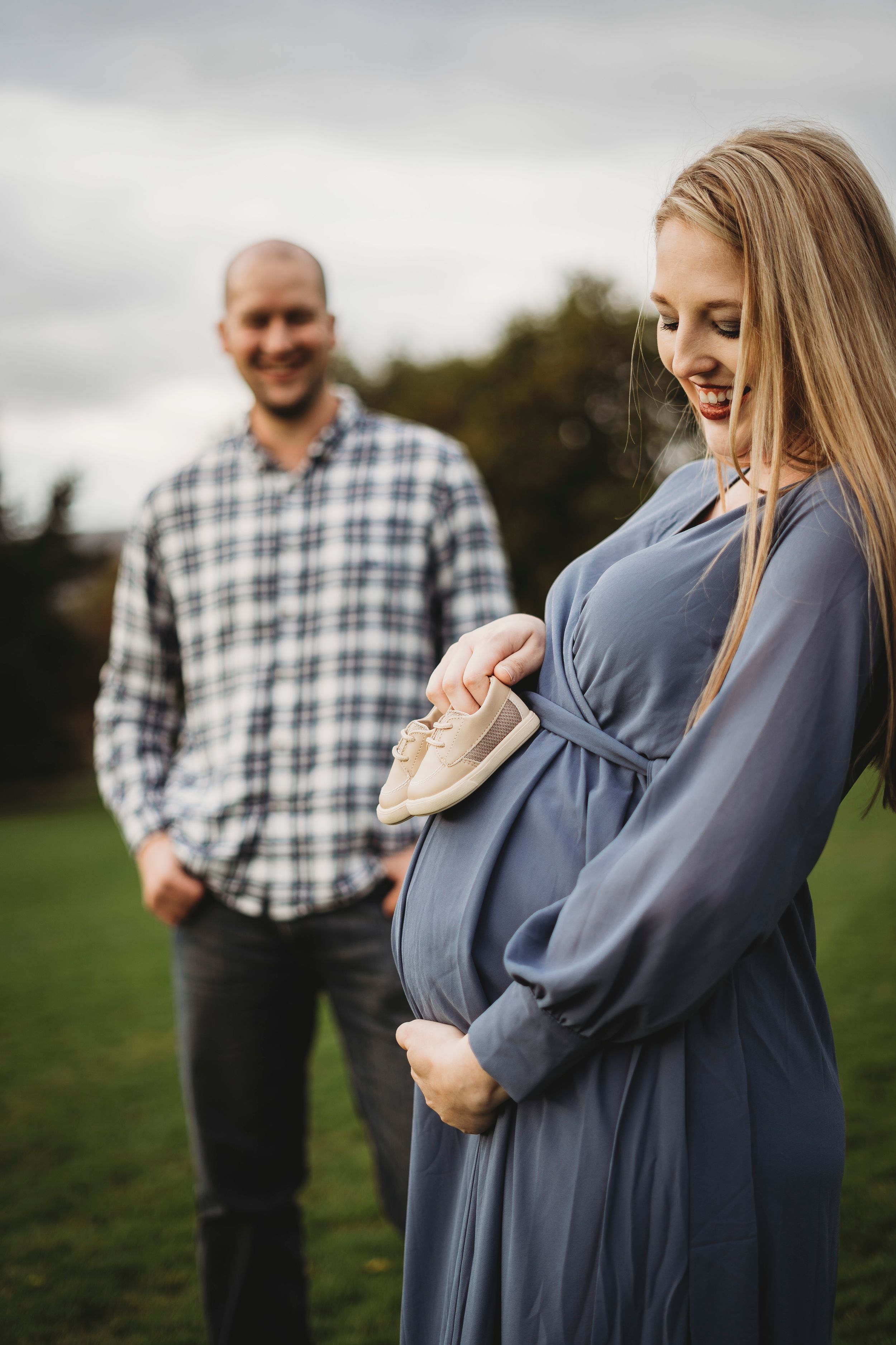 A maternity photo in a field with someone in blue dress and companion in plaid shirt.