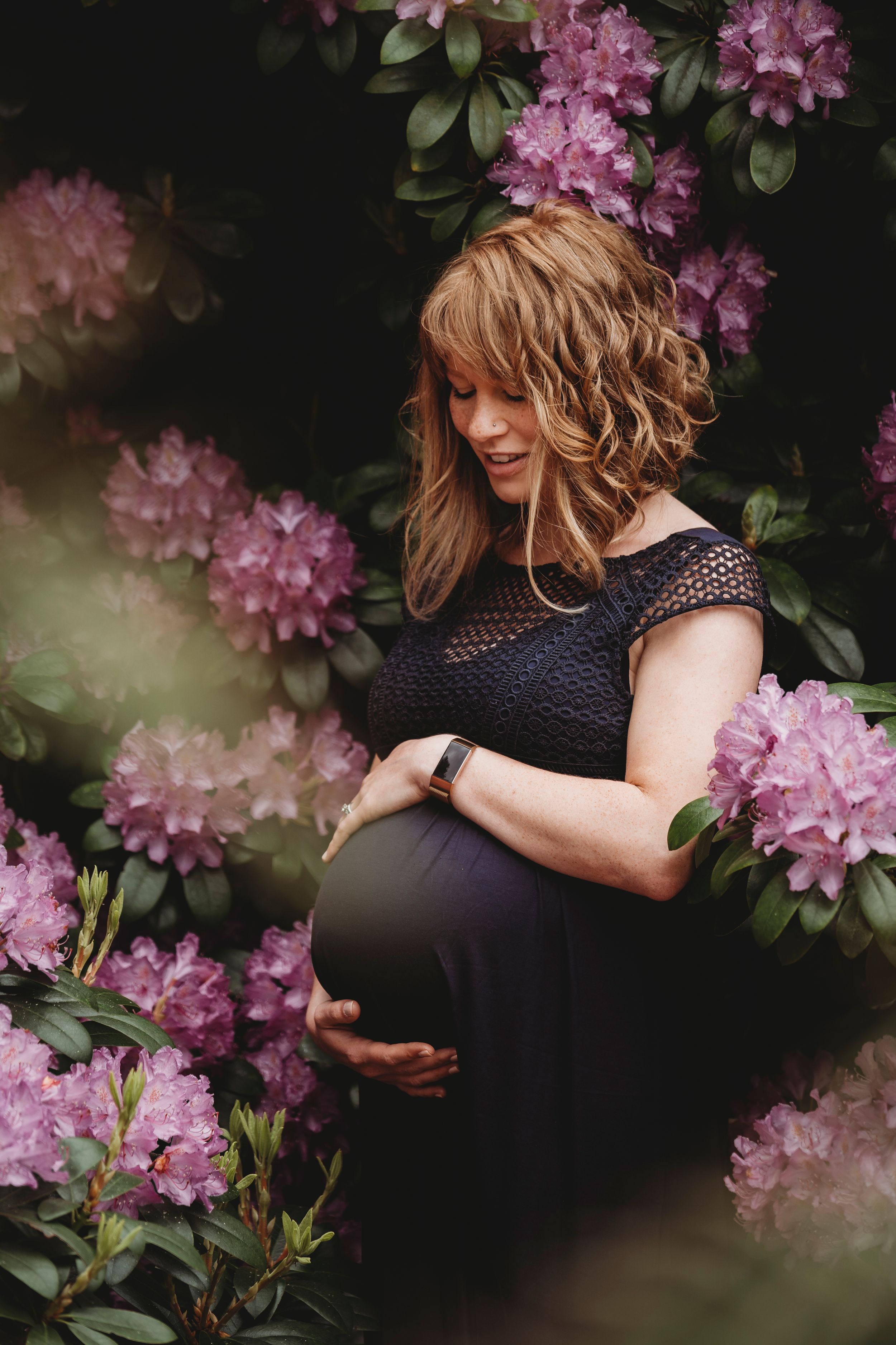 A maternity portrait surrounded by pink flowering bushes in soft lighting.