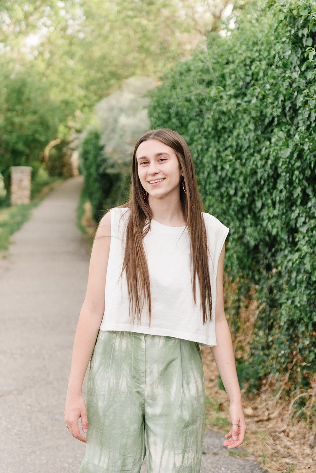 A person in a white crop top and mint green pants stands on a pathway lined with lush greenery.