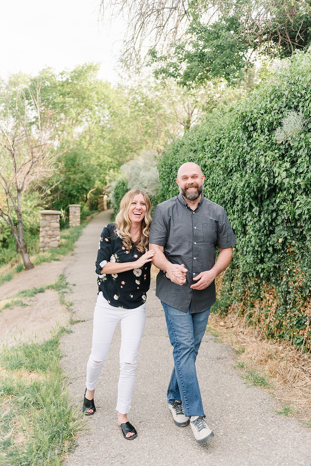 A couple laughing while walking together on a path lined with green hedges and trees.