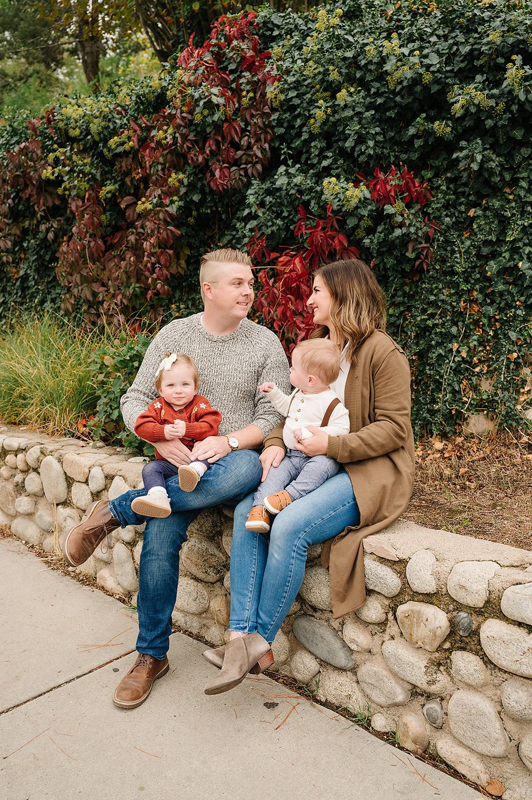 Young family sitting together on a stone wall with colorful fall foliage behind them.