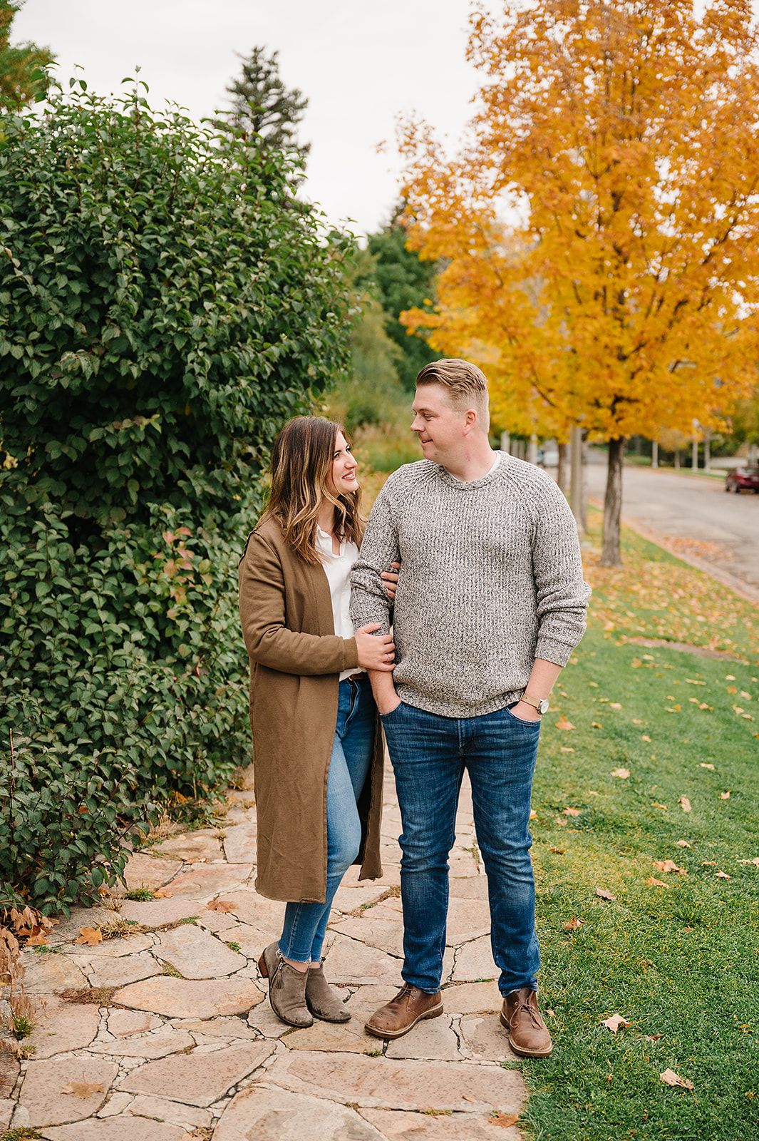 A couple stands together on a stone path surrounded by autumn foliage and green hedges.