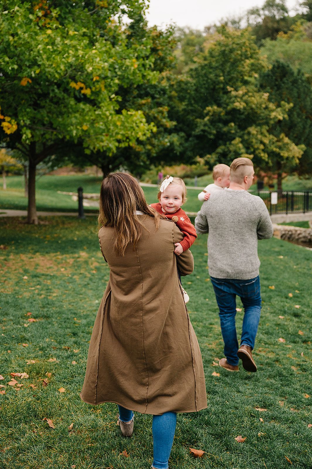 Family walking on grass in a park during fall with colorful trees in the background.