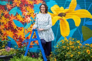 Woman on blue ladder against vibrant floral mural with yellow and orange designs.