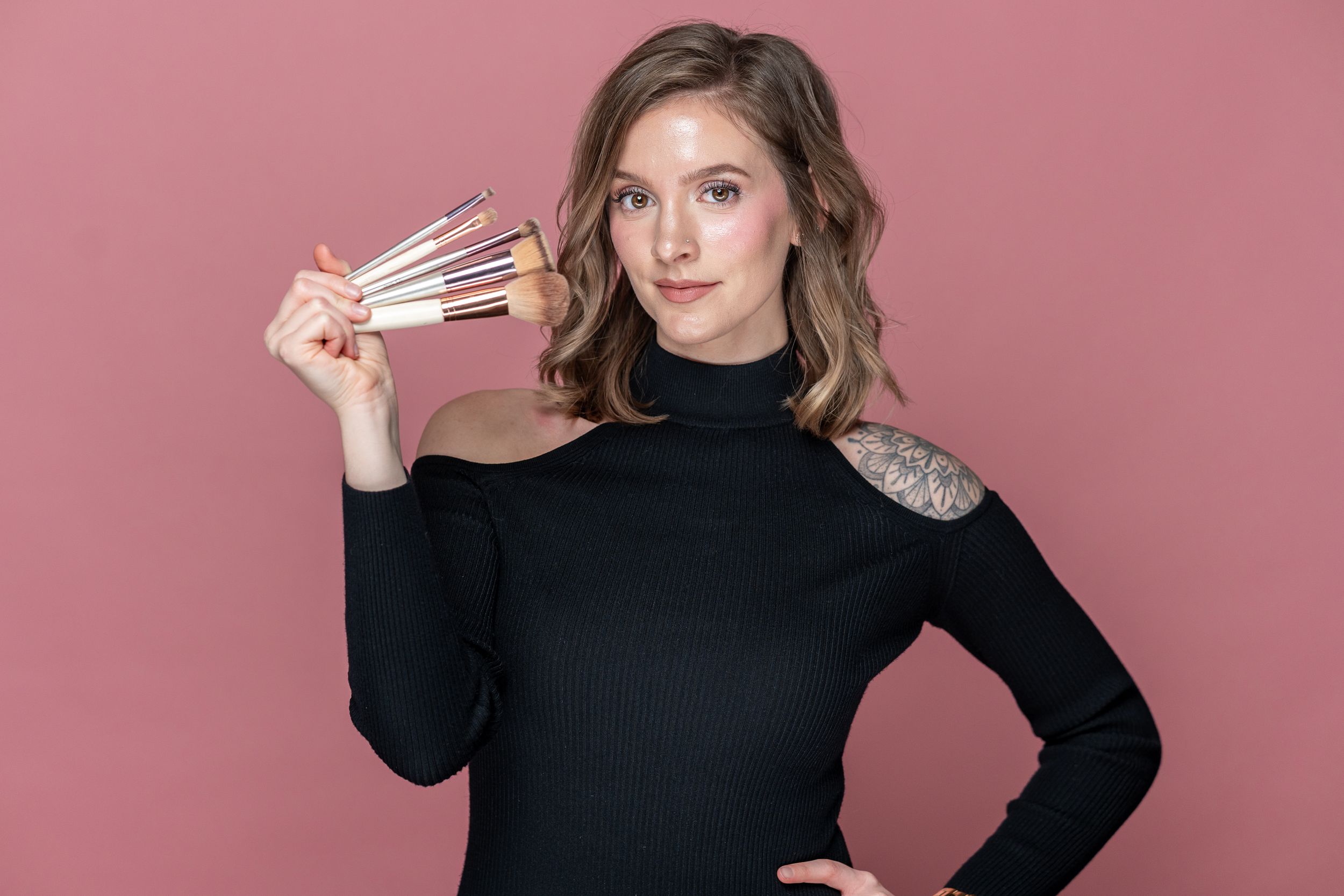 A woman in black holds makeup brushes against a pink backdrop.