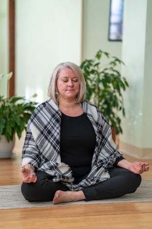 Person in plaid wrap practicing meditation in lotus pose on hardwood floor with plants.
