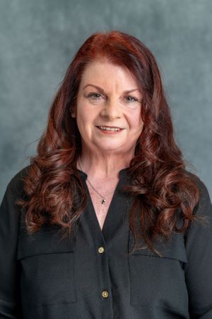 Professional headshot of person with long auburn hair wearing black button-up blouse against gray background.