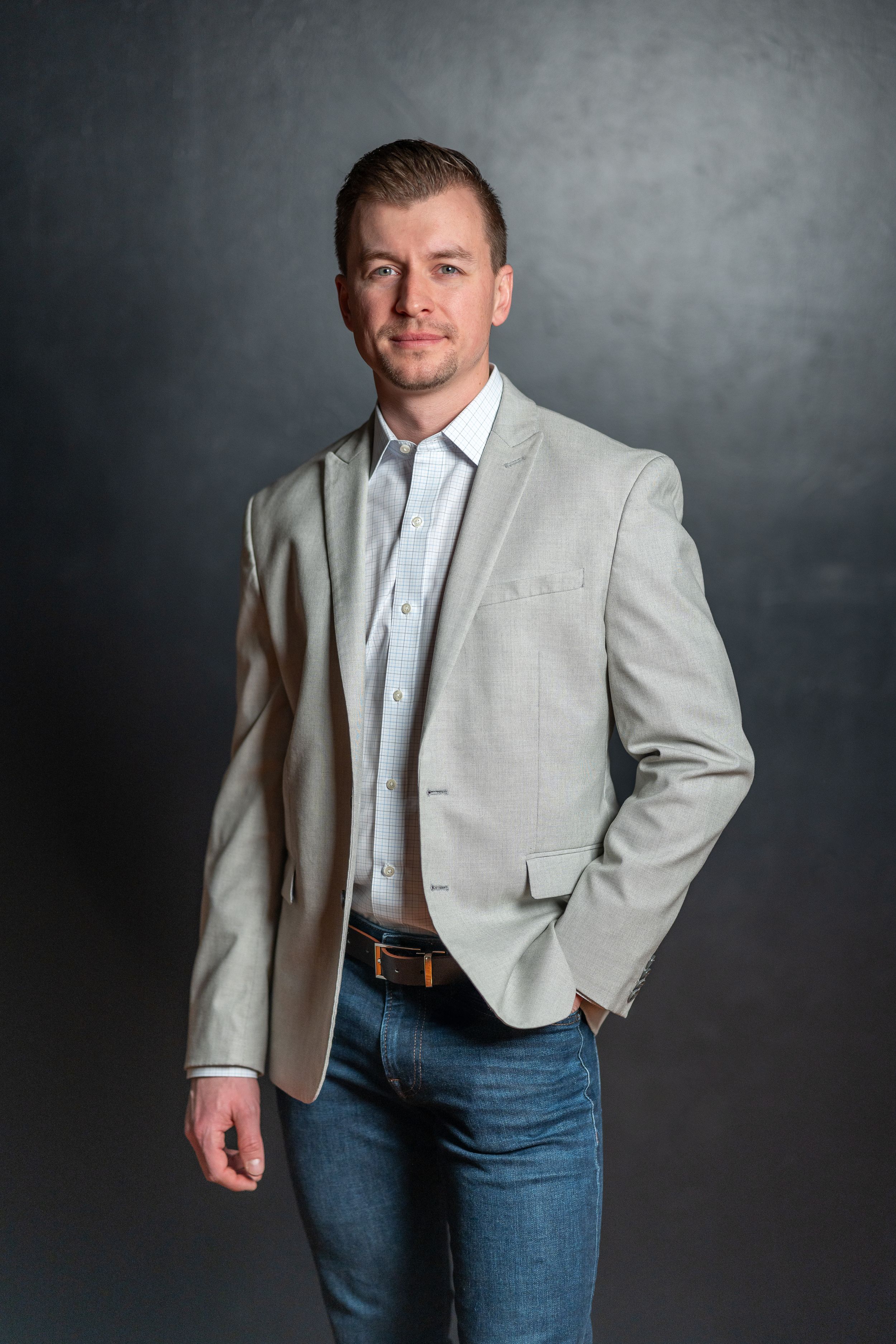 Professional portrait of person in light gray blazer and jeans against dark background.