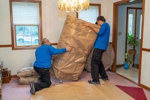 Movers in blue uniforms carefully moving a large wrapped item up carpeted stairs.
