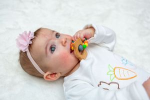 Baby wearing white onesie with pink headband sucking on colorful teether.
