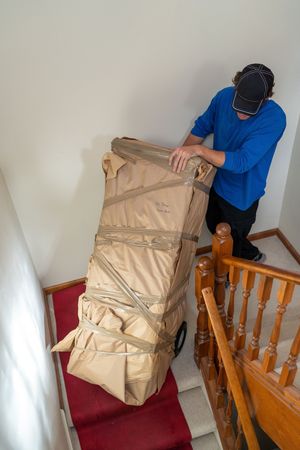 Professional mover handling a wrapped furniture piece on red carpeted stairs.