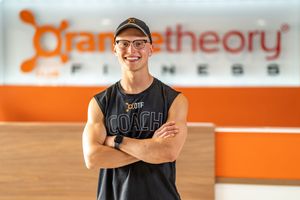 Fitness trainer in black sleeveless shirt poses at Orange Theory gym with logo visible.