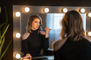 A reflection in a vanity mirror shows someone applying makeup near illuminated bulbs.