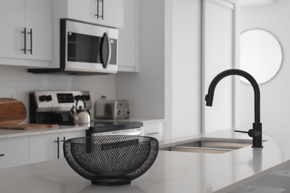 Minimalist white kitchen with matte black faucet and mesh fruit bowl on quartz countertop.