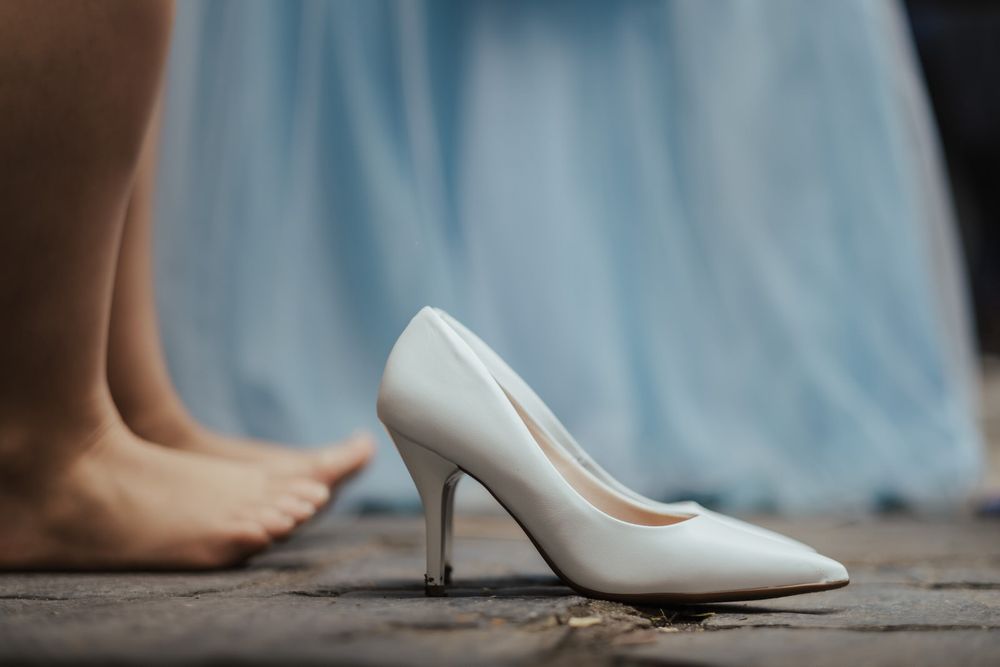 Sapato de salto agulha branco ao lado de pés descalços sobre um fundo de tecido azul claro.