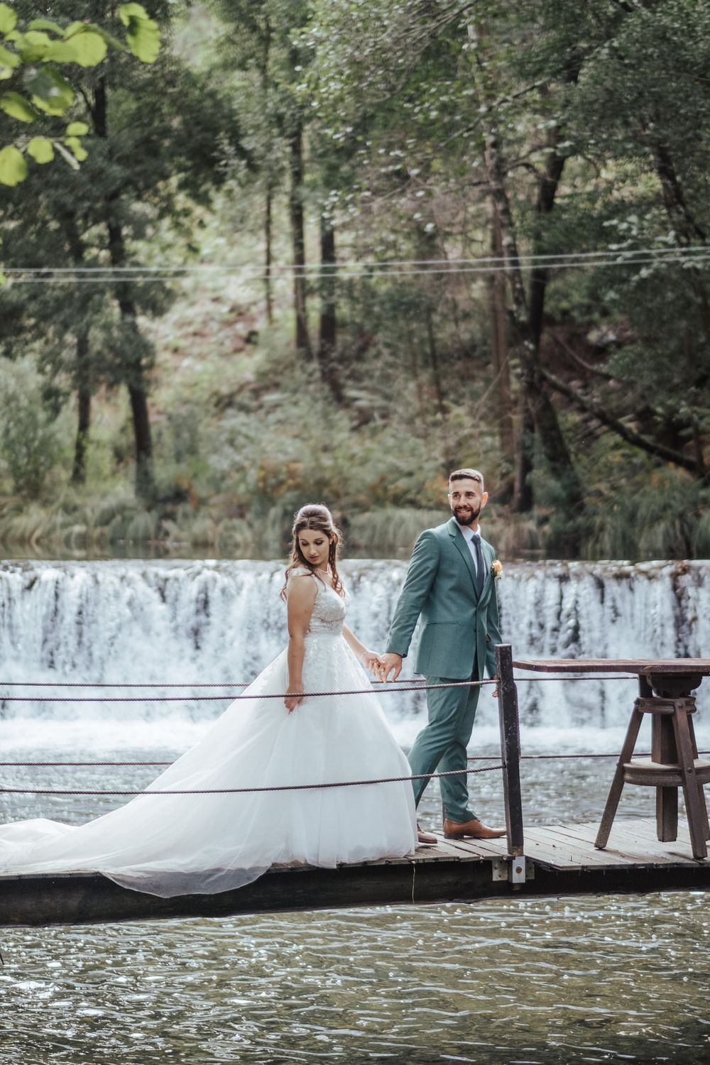 Casal de pé numa ponte de madeira com vista para uma cascata numa zona florestal.