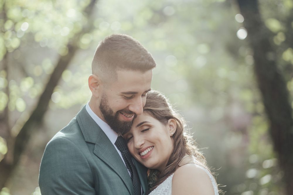 Momento íntimo entre um casal em trajes de casamento, rodeado por uma floresta iluminada pelo sol.