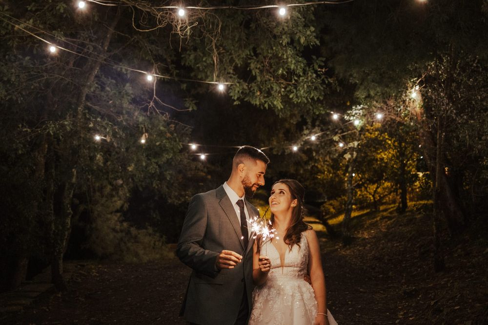 Uma cena romântica de casamento ao entardecer, com luzes de cordão a iluminar um casal debaixo das árvores.