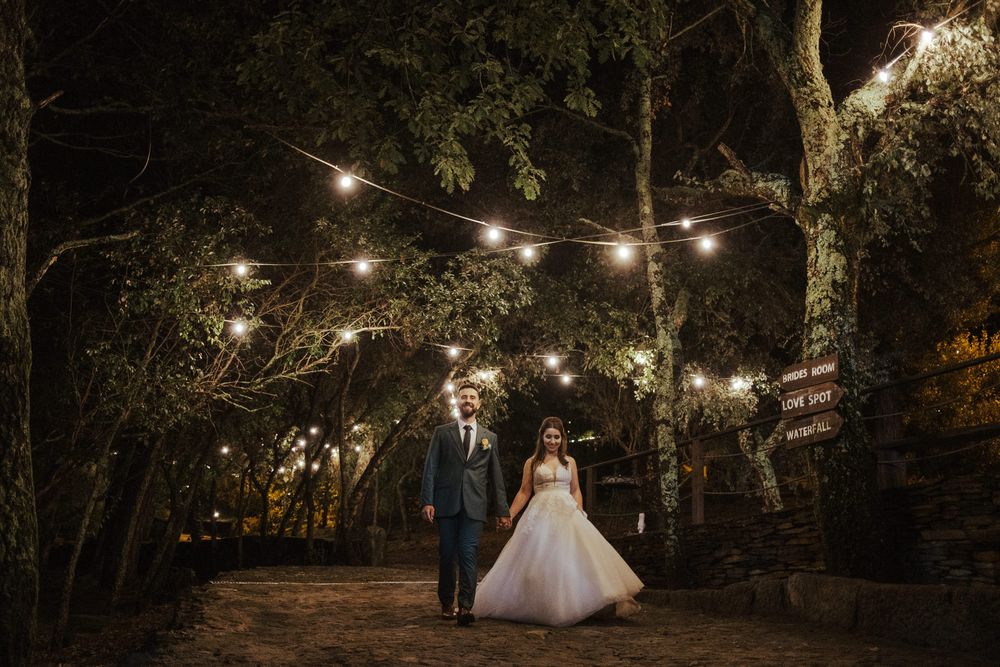 As luzes de cordão iluminam o caminho entre as árvores de um jardim durante um casamento à noite.
