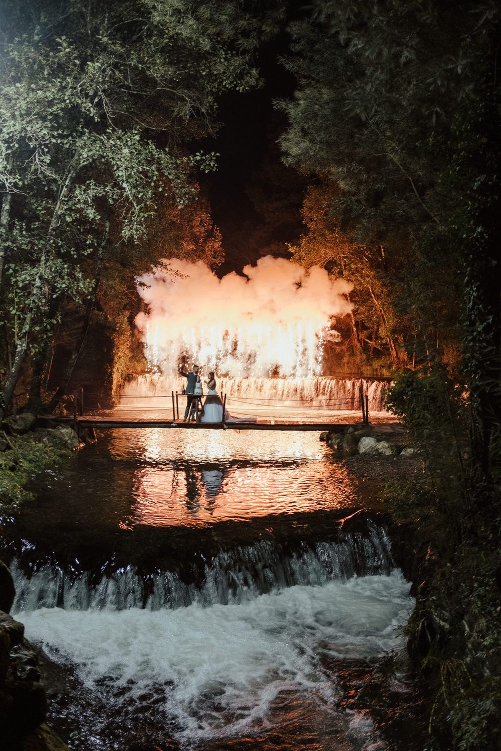 Uma cascata imponente, iluminada por luzes quentes, cria um cenário deslumbrante para um casamento noturno.
