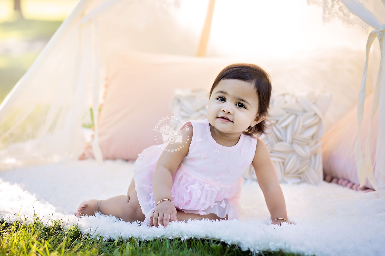 Cute baby girl in pink dress crawling on white fur rug near a teepee outdoors.