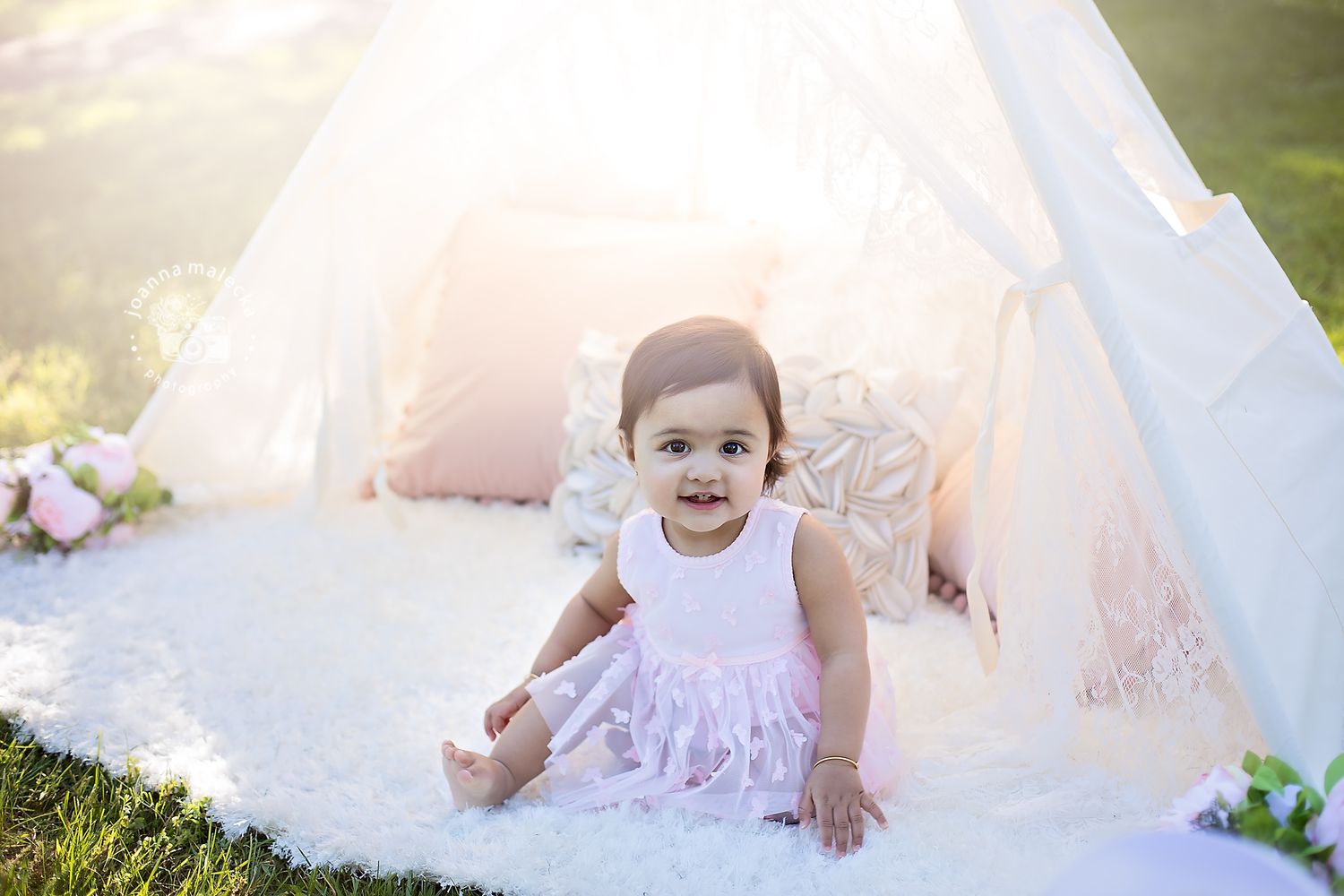Happy baby girl in pink dress sitting on white rug in front of a white teepee.