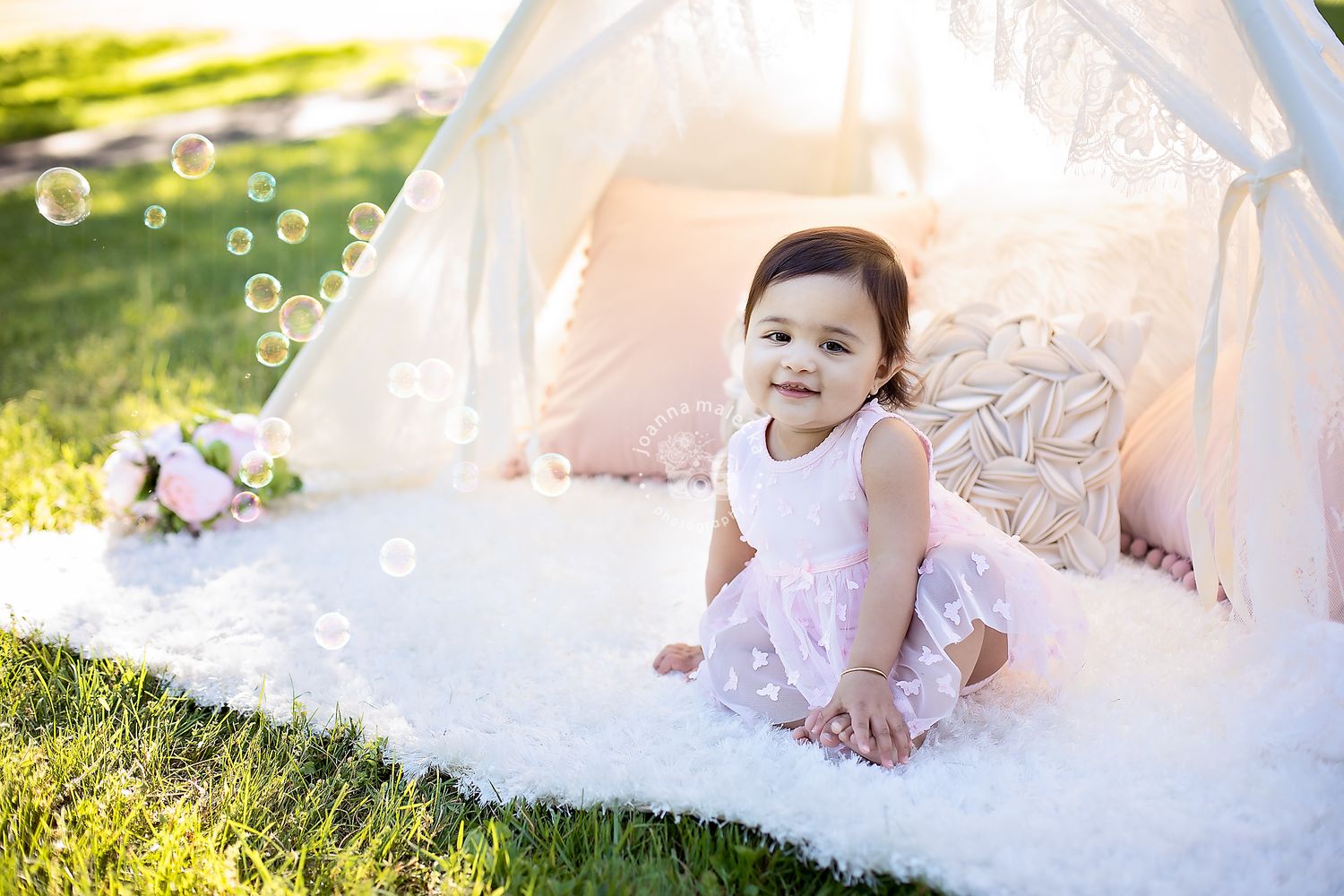 Smiling baby girl in pink dress sitting on white fur rug near teepee tent with bubbles floating around her.