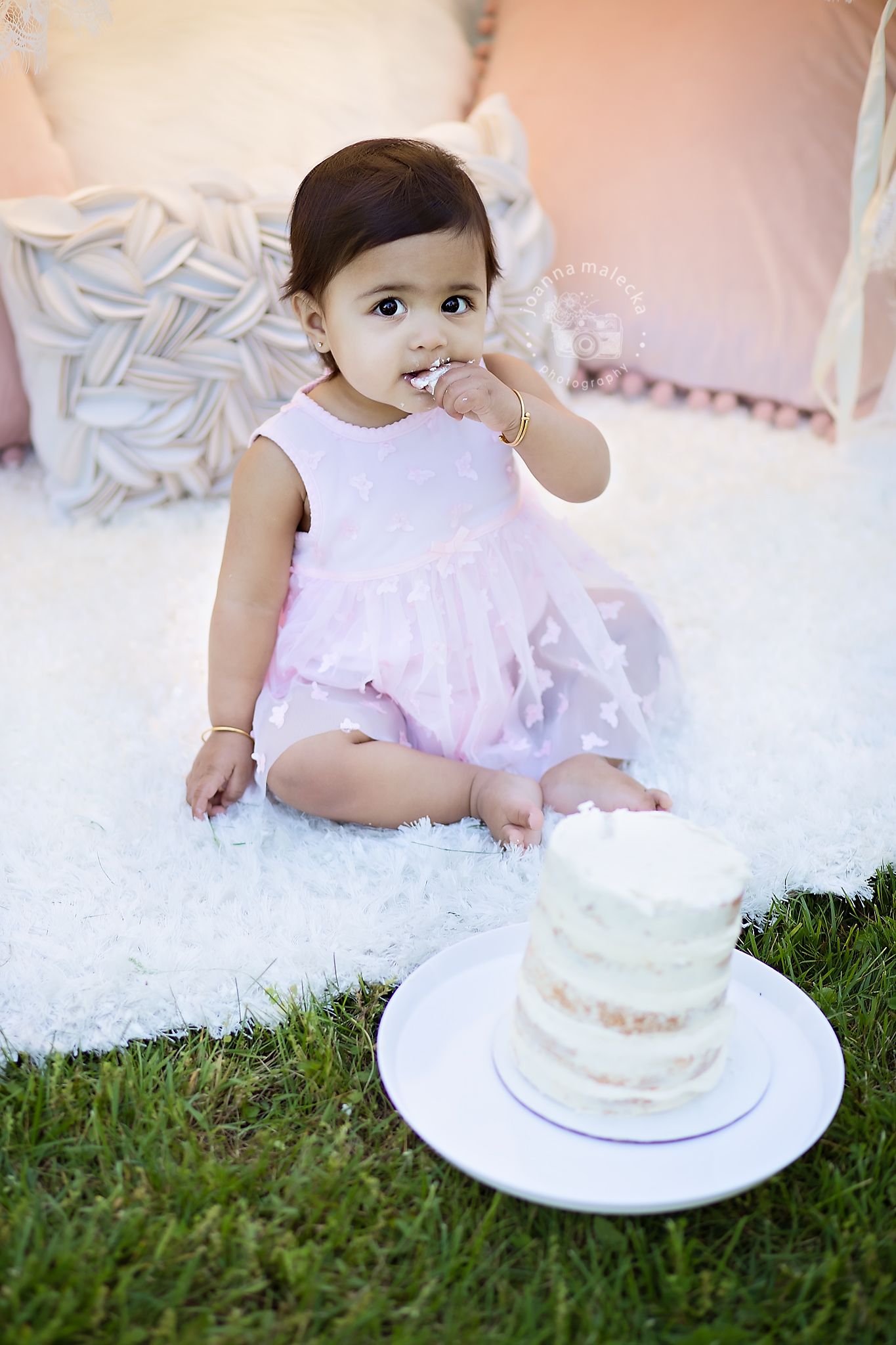 Adorable baby girl in pink tutu dress tasting her first birthday smash cake outdoors on white rug.