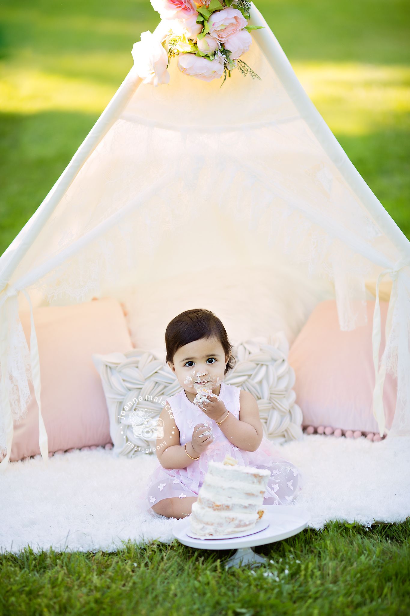 Baby girl in pink dress smashing her first birthday cake in front of a white floral teepee tent.