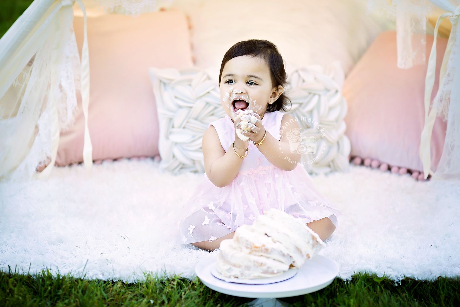 Joyful baby girl in pink dress laughing while smashing birthday cake outdoors near white teepee tent.