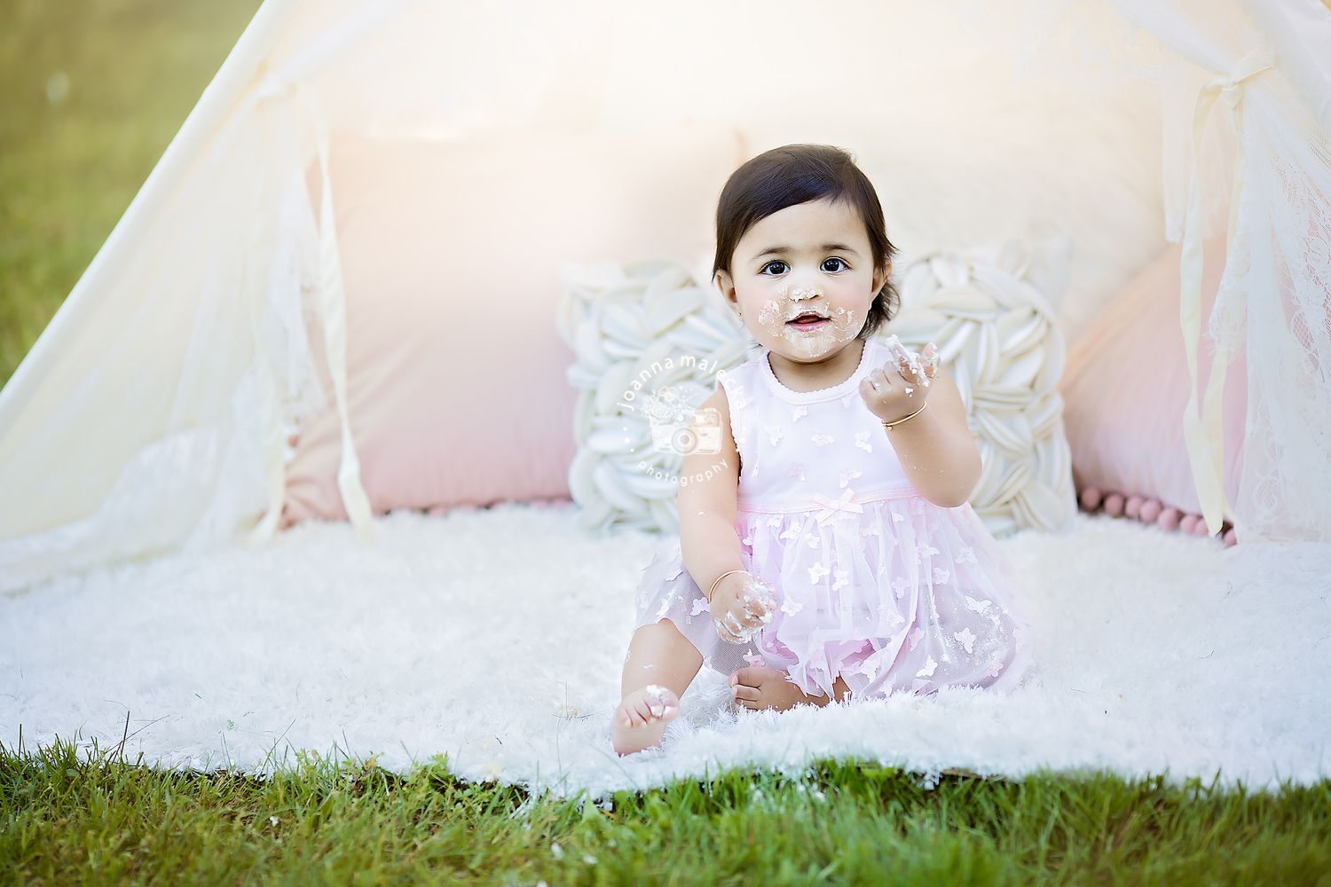Happy baby girl in pink dress sitting on white fur rug in front of glowing white teepee tent.