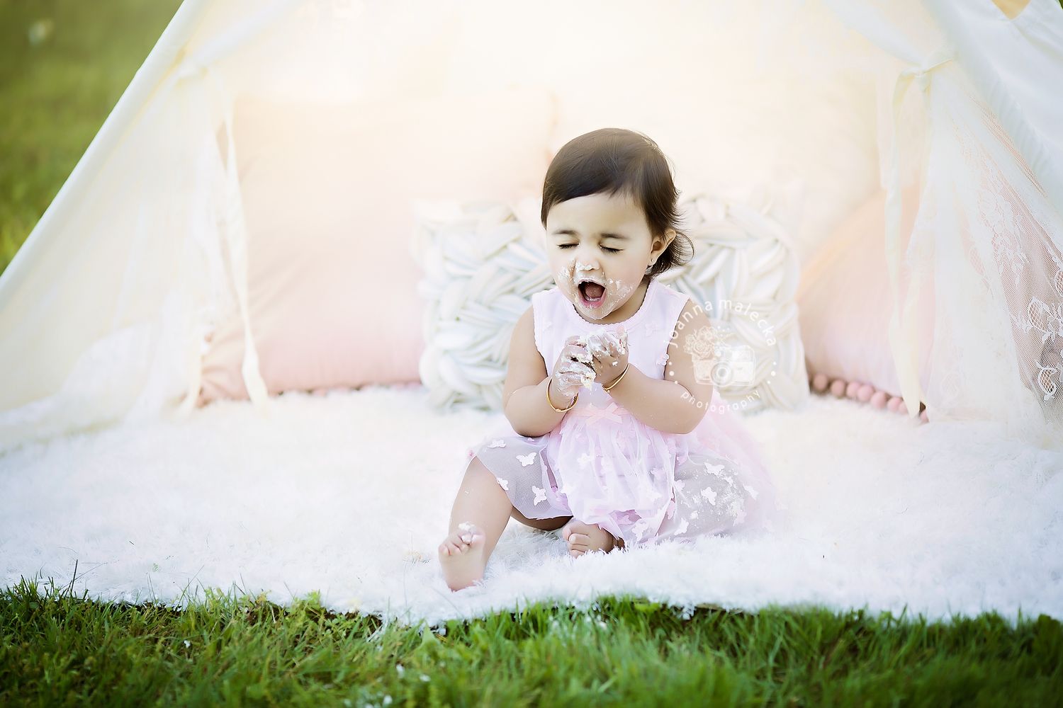 Cute baby girl in pink dress looking down at cake crumbs while sitting near white teepee tent.