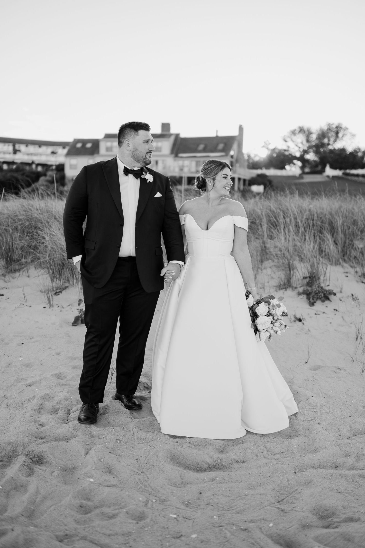 Wedding couple walks along sandy beach dunes with beach houses in the distance.