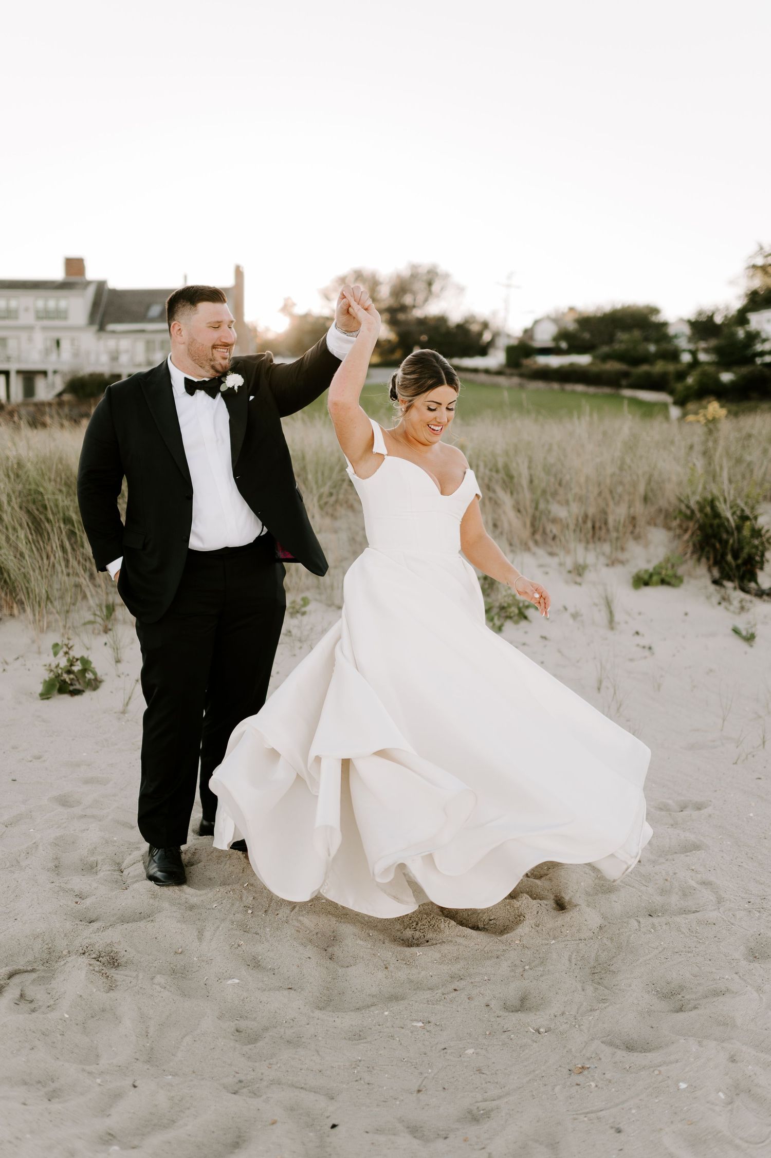 Bride's white dress flows in the wind as couple dances on beach dunes during sunset.