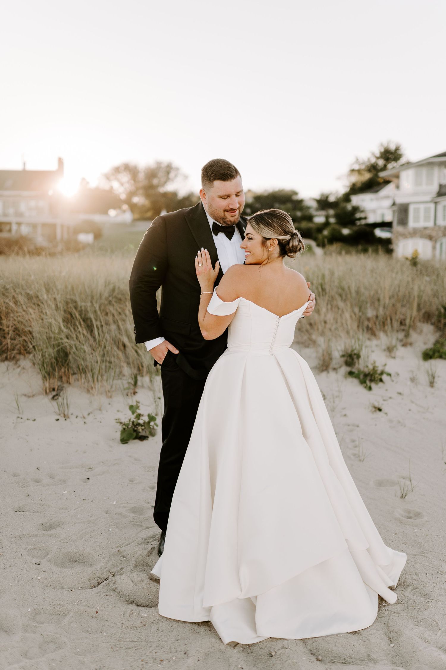 Newlyweds share candid moments on a beach with coastal houses visible in the background.