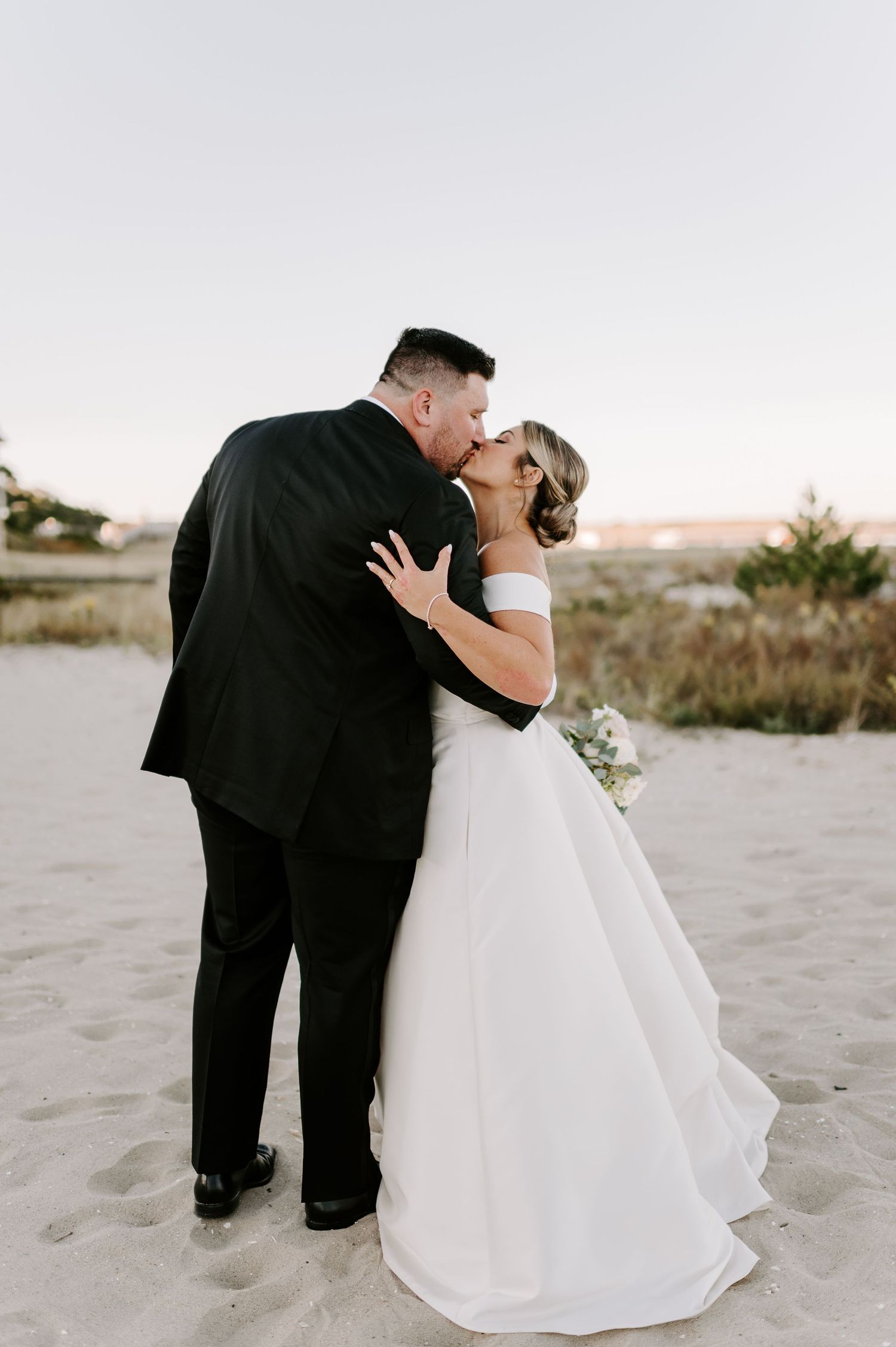 Newlyweds embrace playfully on a beach with dunes and coastal vegetation in the background.