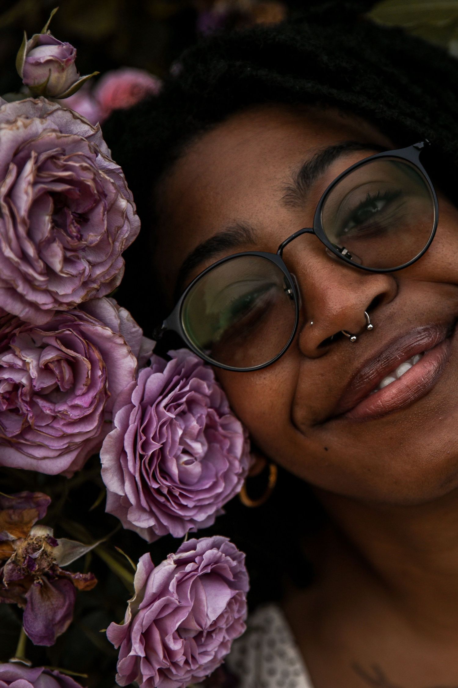 Dark-skinned girl with dark hair and glasses smiles, leaning into purple roses.