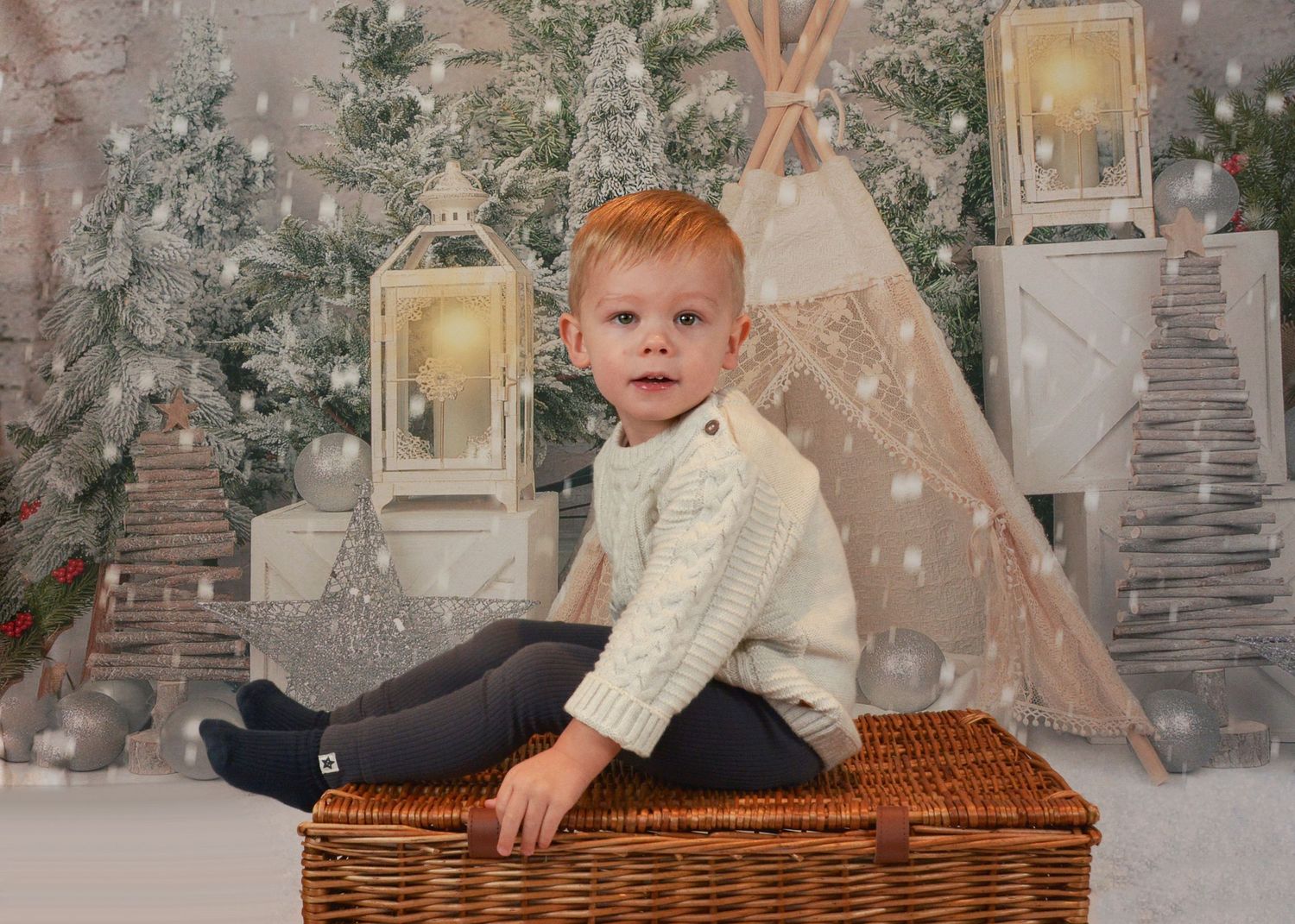 Young toddler boy sitting on a wicker basket in a festive Christmas studio setting.