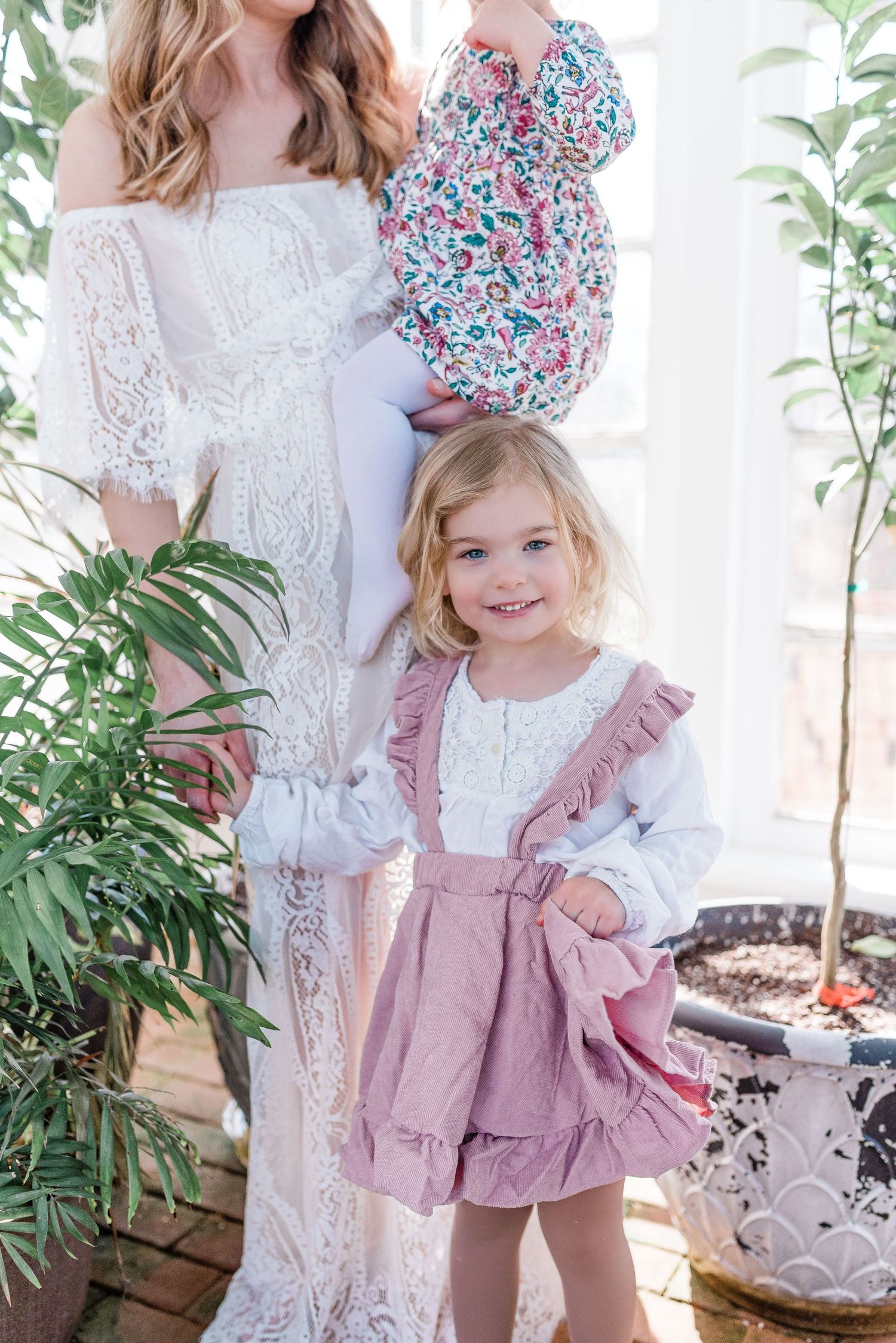 White lace dress and child in pink ruffle romper pose together among potted plants in a bright indoor setting.