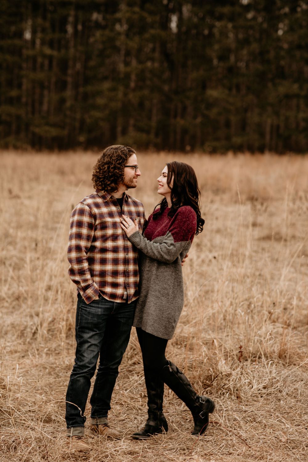 Couple standing together in dry autumn meadow with pine forest backdrop, sharing a loving glance at each other.