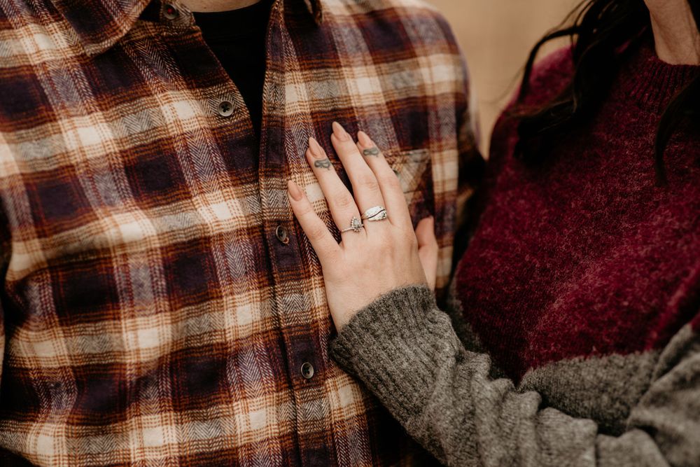 Close-up of womans hand with engagement ring resting on mans brown plaid flannel shirt in autumn setting.