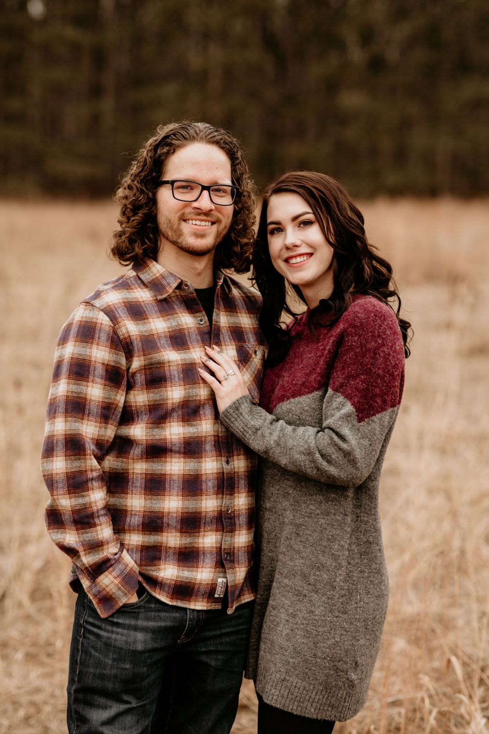 Smiling engaged couple posing in autumn field, man in plaid shirt and woman in gray burgundy sweater looking at camera.