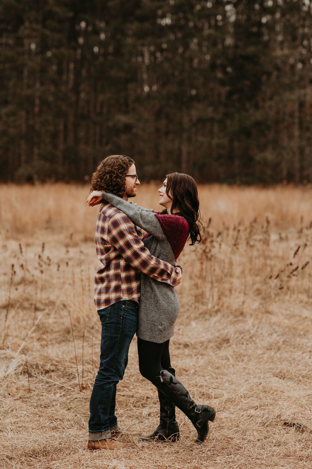 Couple embracing in a golden field with pine trees, wearing flannel and gray sweater in autumn.