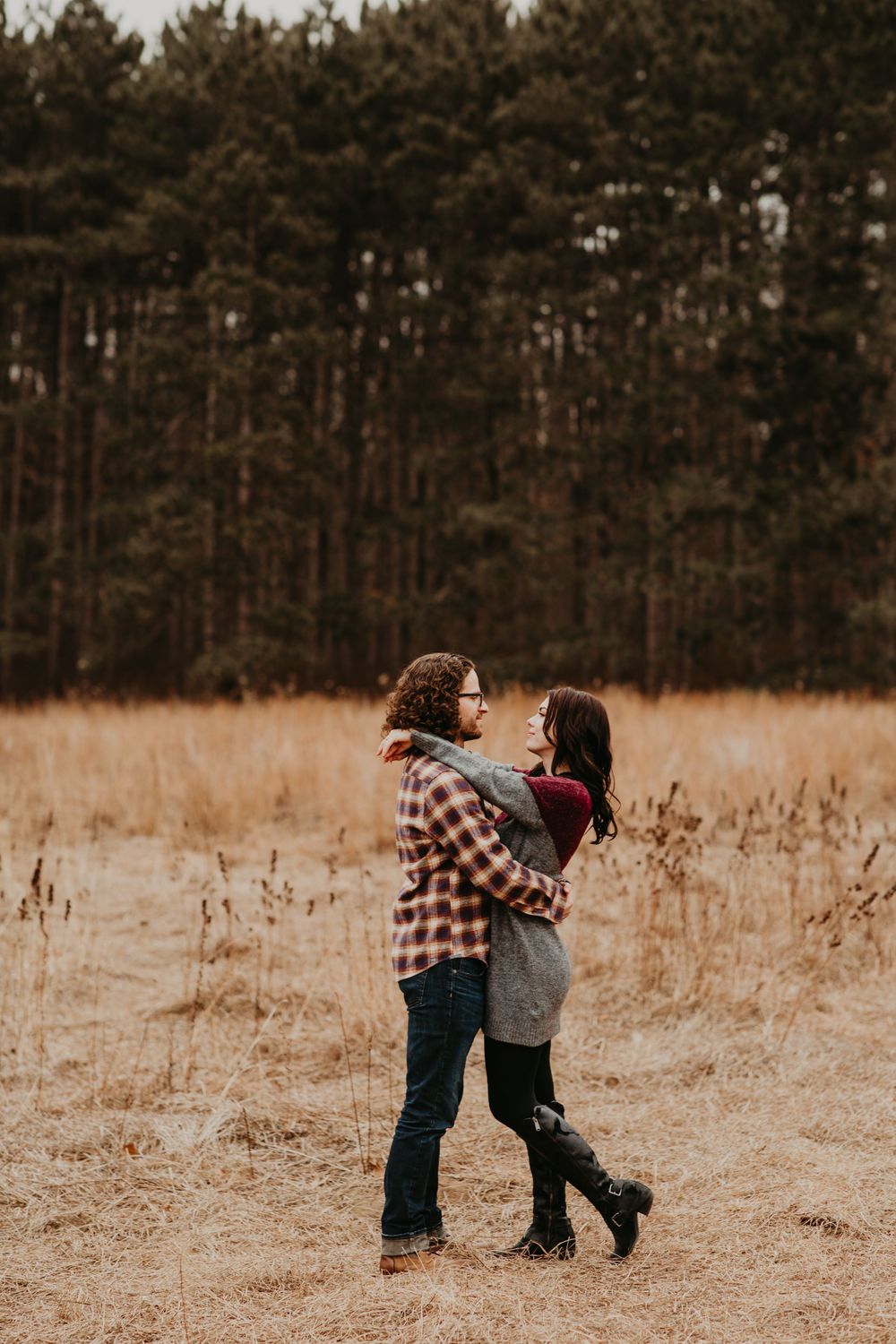 Romantic couple holding each other close in a dry autumn field surrounded by tall pine trees.