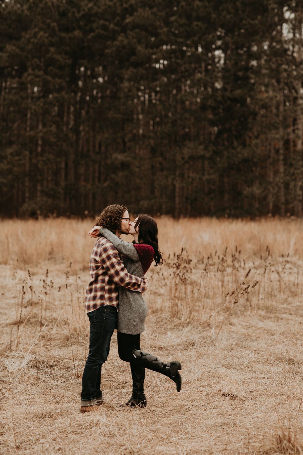 Couple sharing a kiss in a golden meadow with pine forest backdrop during autumn season.