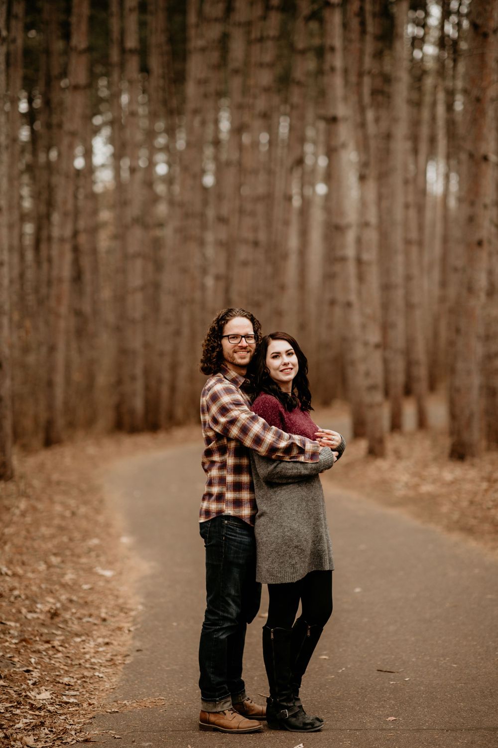 Couple hugging on a tree-lined path in autumn, man in flannel shirt embracing woman in gray sweater.