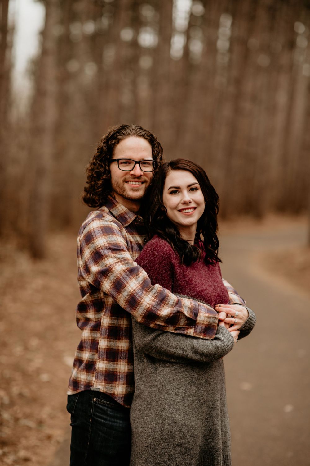 Smiling couple embracing on a forest path in fall, man in plaid shirt holding woman in burgundy sweater.
